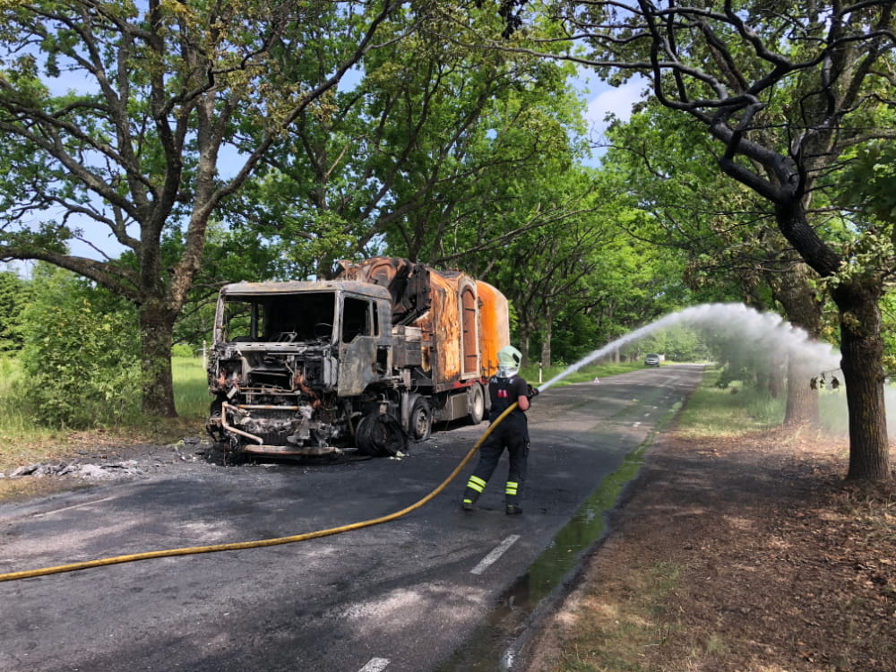 Firefighter hosing down a burnt-out lorry carrying a Revonia concrete module on a tree-lined road